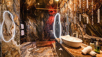 Badezimmer der Juniorsuite Kuscheltraum Deluxe Luxuriöser Waschtischbereich der Juniorsuite Kuscheltraum Deluxe im Hotel Winzer mit Naturstein, beleuchtetem Spiegel und Kristalldetails