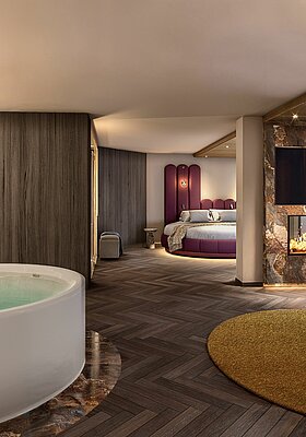 Visualisierung einer luxuriösen Hotel-Wellness-Suite. Im Vordergrund steht eine große, freistehende Whirlpool-Badewanne auf einem marmorierten Sockel. Rechts daneben befindet sich ein gemütlicher Sitzbereich mit einem runden Loungesessel, einem Beistelltisch und einem Kamin unter einem Fernseher. Im Hintergrund ist ein großer kreisförmiger Schlafbereich mit einem markanten violetten Kopfteil zu sehen. Die Suite zeichnet sich durch dunkles Fischgrätparkett, strukturierte Holz- und Marmorwände sowie warme, gedimmte Beleuchtung aus.